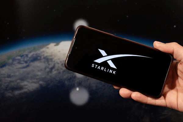 มือที่กำลังโทรศัพท์ที่หน้าจอฉายโลโก้  Starlink พร้อมพื้นหลังเป็นรูปโลกและอวกาศ 