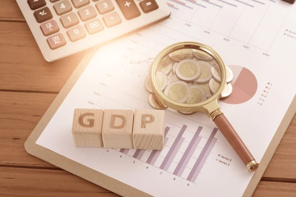 บล็อคไม้ต่อกันเป็นคำว่า GDP และมีแว่นขยาย เหรียญเงิน พร้อมกราฟแสดงข้อมูล สื่อถึงการนำค่า GDP มาวิเคราะห์ตลาด 