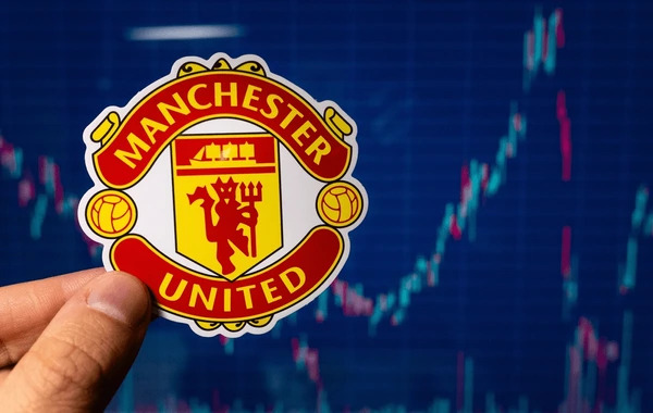 โลโก้ของสโมสรฟุตบอลชื่อดัง Manchester United กำลังถูกถือโดยมีกราฟราคาหุ้นอยู่ด้านหลัง สื่อถึงความผันผวนของราคาหุ้นสโมสรฟุตบอล 